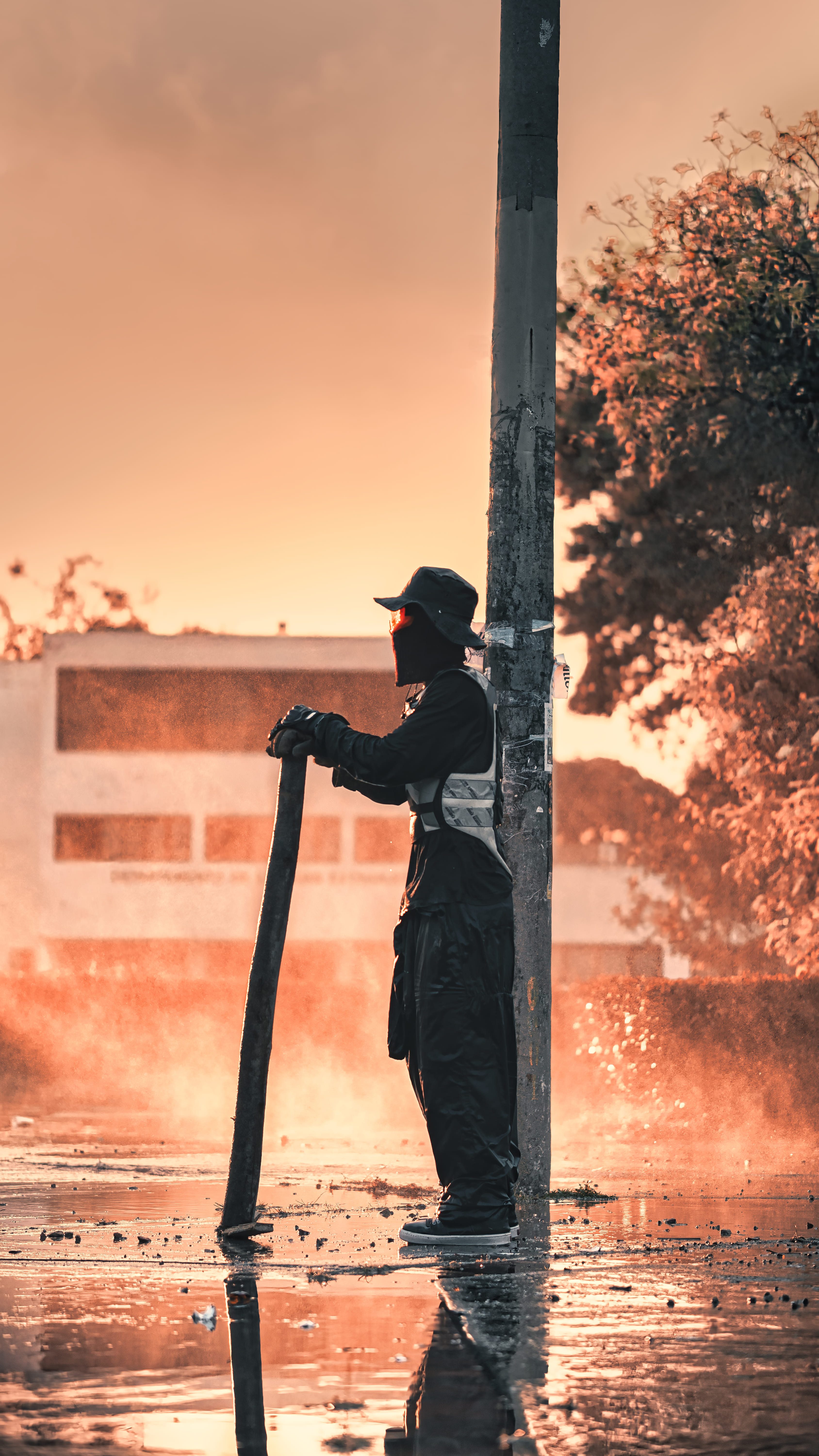 Hombre con manguera apoyado en un poste durante un atardecer con agua y humo